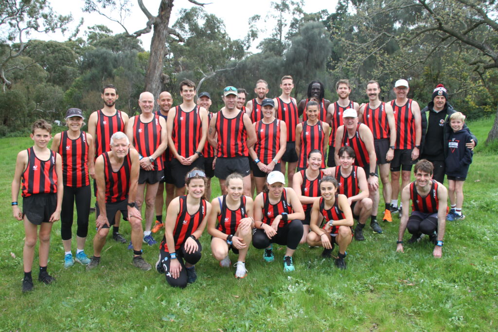 A group of runners standing together in red and black striped shirts. The group stands on grass in front of trees.
